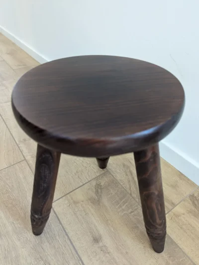 Petit tabouret tripode en bois foncé avec assise ronde et pieds sculptés de style brutaliste ou rustique.