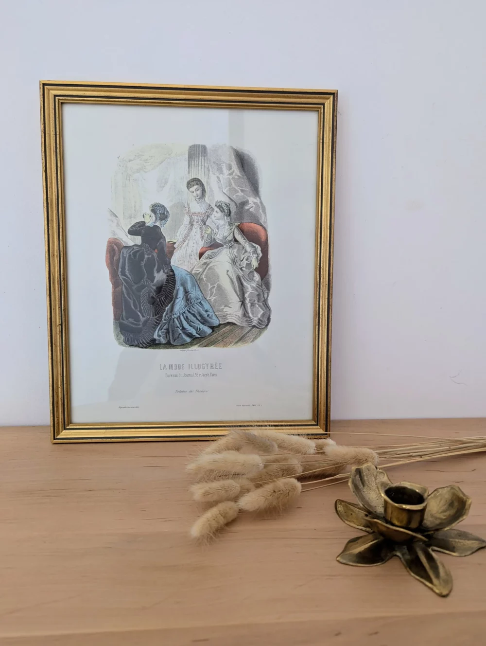 Lithographie "La mode illustrée" posée sur une console avec un bougeoir en laiton et quelques fleurs séchées.