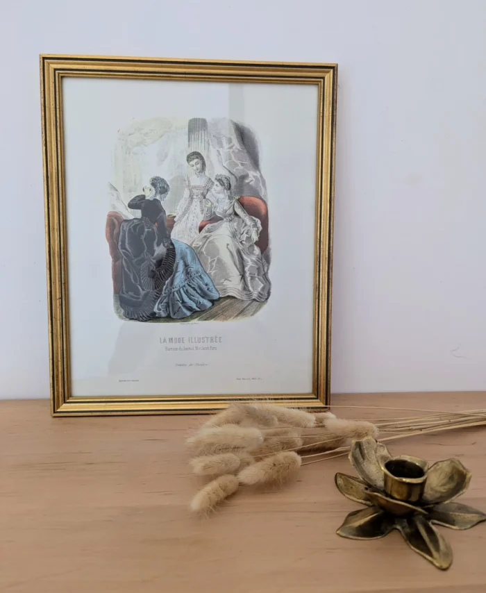 Lithographie "La mode illustrée" posée sur une console avec un bougeoir en laiton et quelques fleurs séchées.