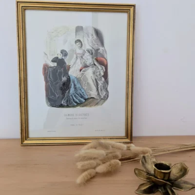 Lithographie "La mode illustrée" posée sur une console avec un bougeoir en laiton et quelques fleurs séchées.