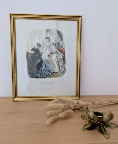 Lithographie "La mode illustrée" posée sur une console avec un bougeoir en laiton et quelques fleurs séchées.