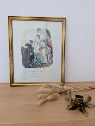 Lithographie "La mode illustrée" posée sur une console avec un bougeoir en laiton et quelques fleurs séchées.