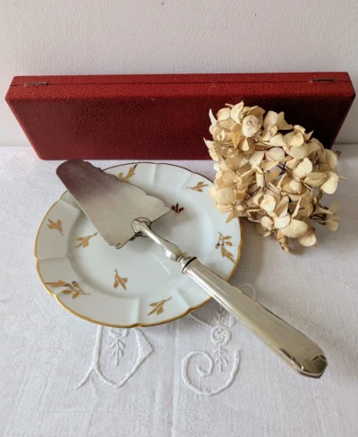 Pelle à tarte vintage en métal blanc posée sur une assiette ancienne avec son écrin rouge en arrière-plan