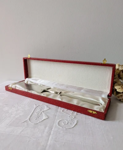 Pelle à tarte en métal blanc rangée dans son coffret rouge d’origine sur nappe brodée