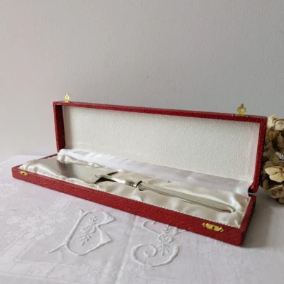 Pelle à tarte en métal blanc rangée dans son coffret rouge d’origine sur nappe brodée