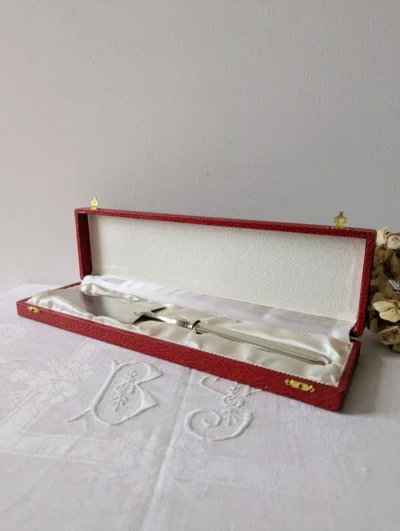 Pelle à tarte en métal blanc rangée dans son coffret rouge d’origine sur nappe brodée