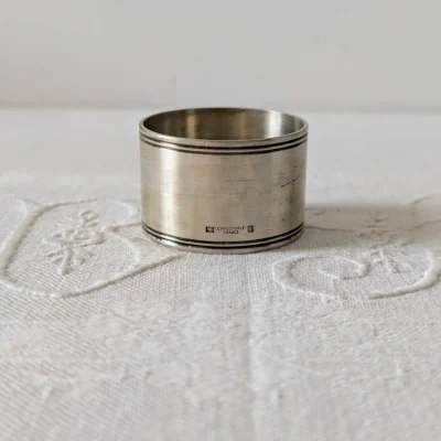 Rond de serviette Christofle ancien en métal argenté posé sur nappe blanche.