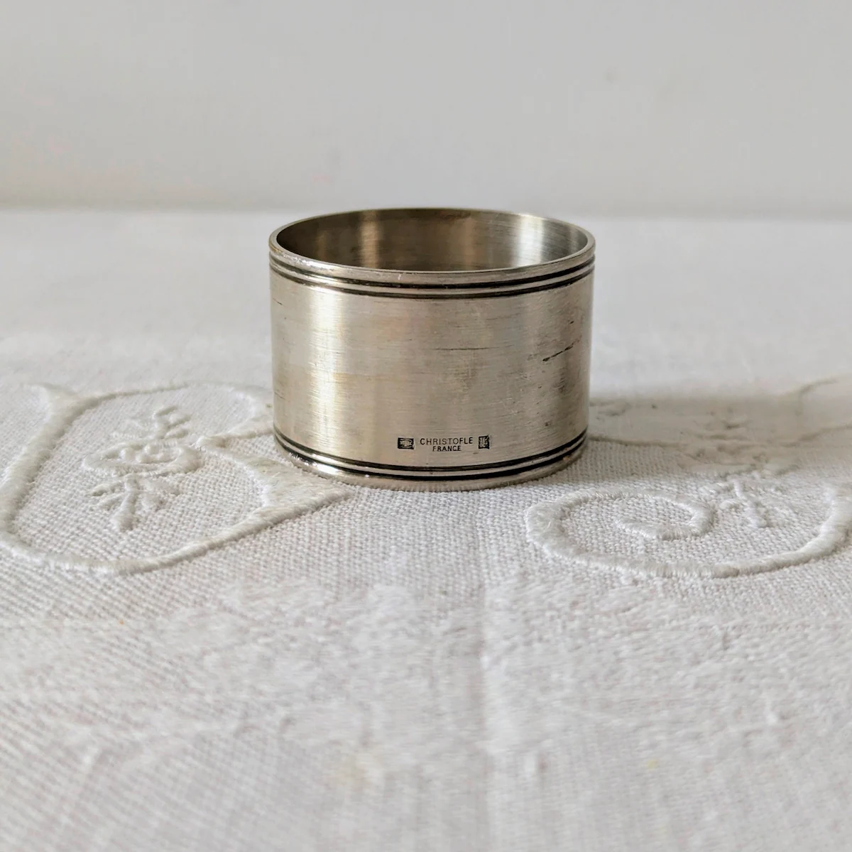 Rond de serviette Christofle ancien en métal argenté posé sur nappe blanche.