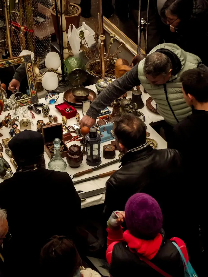 Vue plongeante sur une table de brocanteur présentant une collection d'objets d'art, verreries et bibelots scrutés par des chineurs avertis.