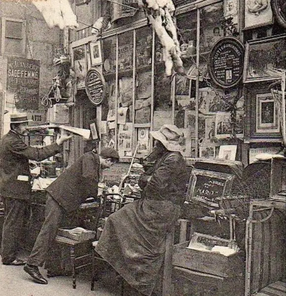 Photographie ancienne montrant un chineur examinent avec attention un objet sur l'étal encombré d'un brocanteur parisien au début du XXe siècle.