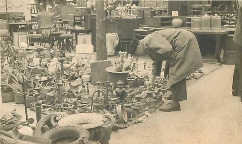 Chineur scrutant un déballage d'objets anciens au sol aux Puces de Saint-Ouen au milieu du XXe siècle.