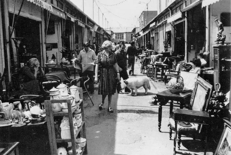 Scène de vie dans une allée pavée des Puces de Saint-Ouen au XXe siècle, illustrant la professionnalisation des stands d'antiquités.