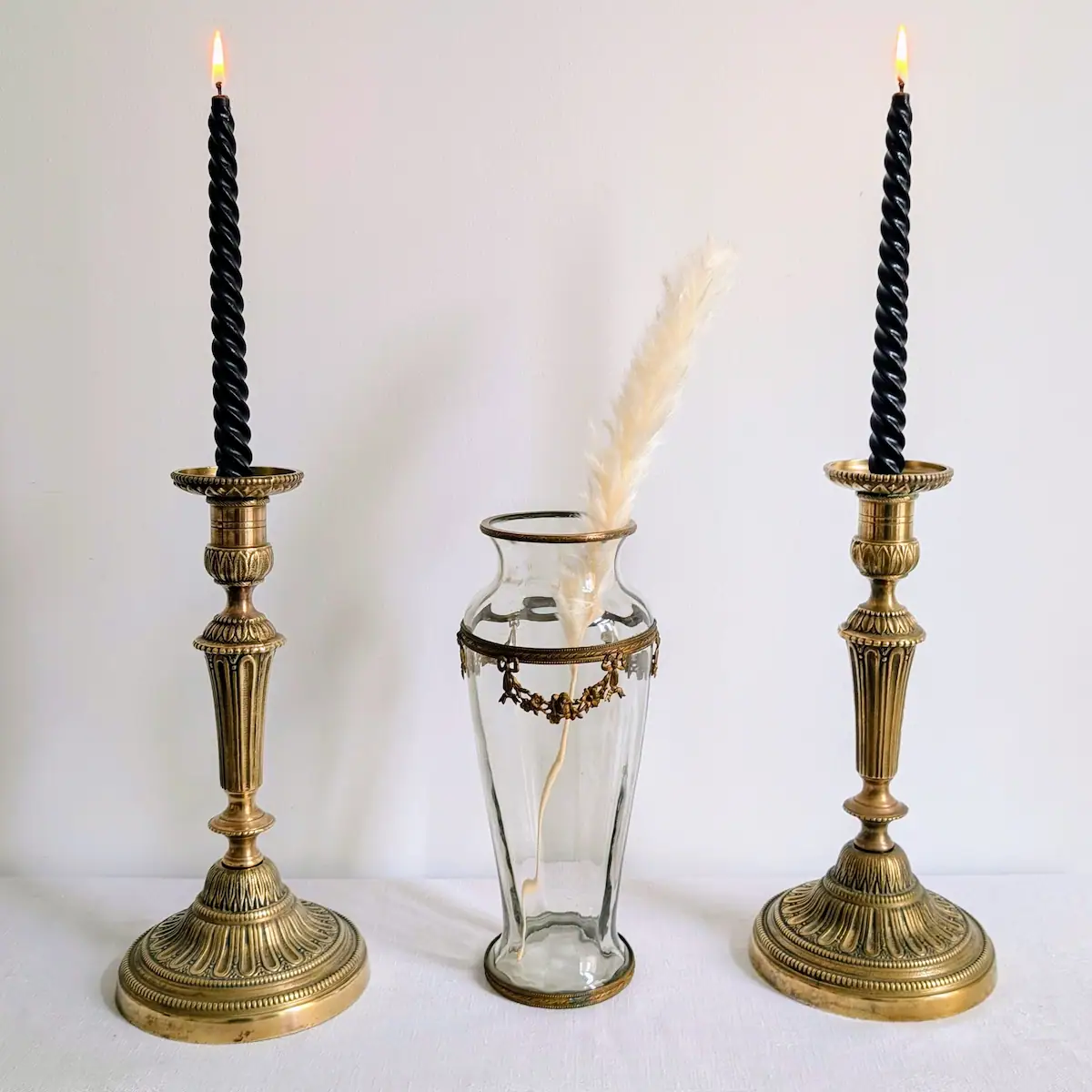 Flambeaux en laiton de style Louis XVI allumés avec cierges noirs et vase en verre transparent.