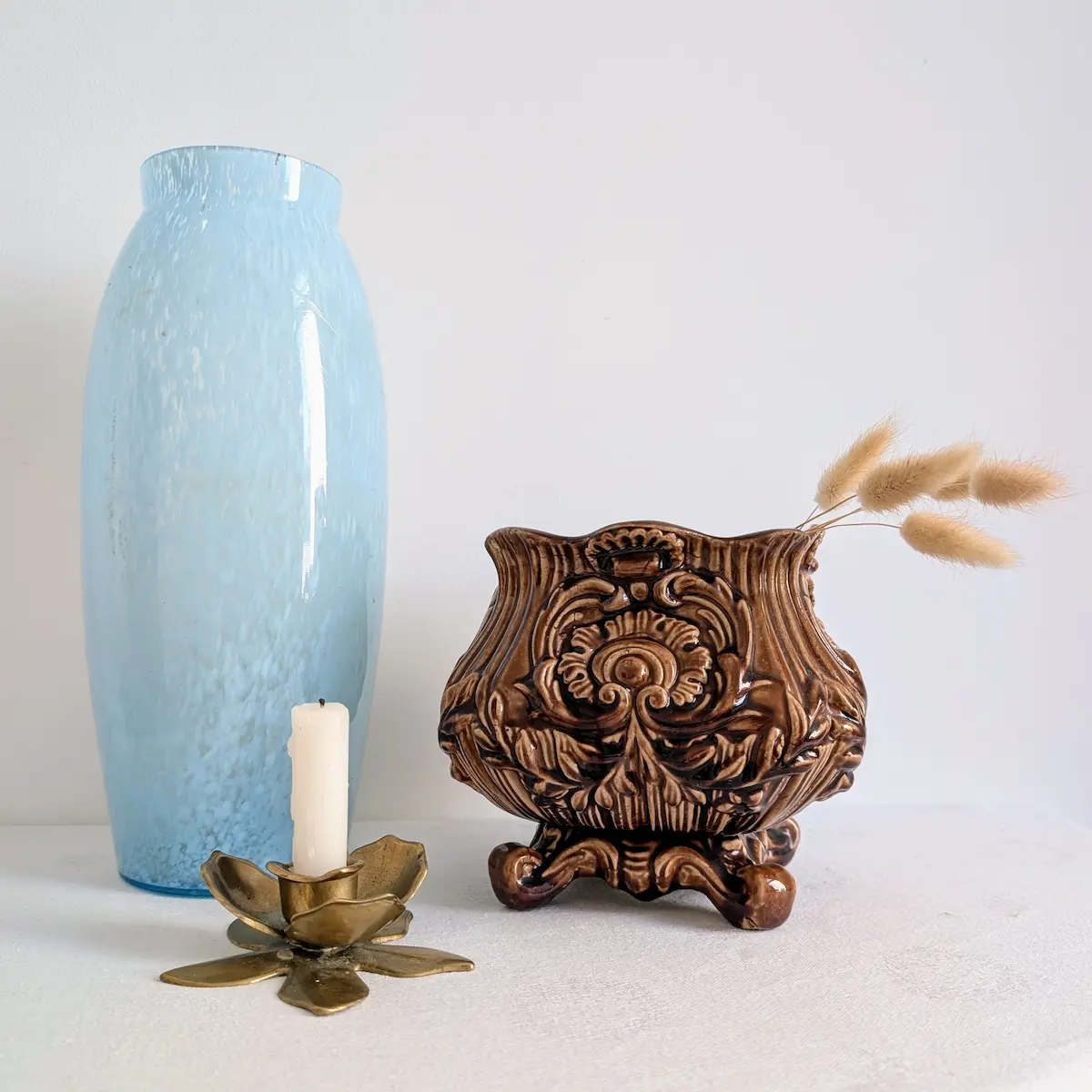 Mise en scène d'un cache-pot en barbotine brune à côté d'un vase en verre et d'un bougeoir en laiton.