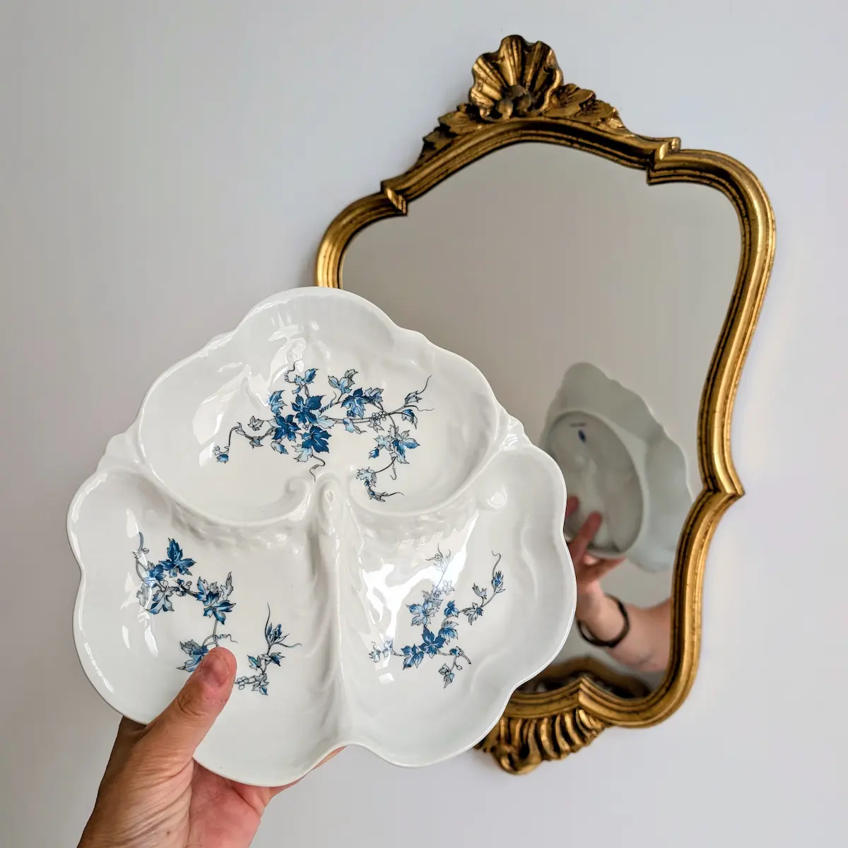 Main tenant un serviteur en porcelaine Bernardaud devant un miroir vintage en bois doré