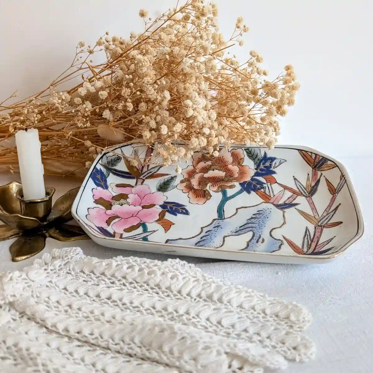 Plateau décoratif en porcelaine posé sur une nappe blanche avec des fleurs séchées et un bougeoir.