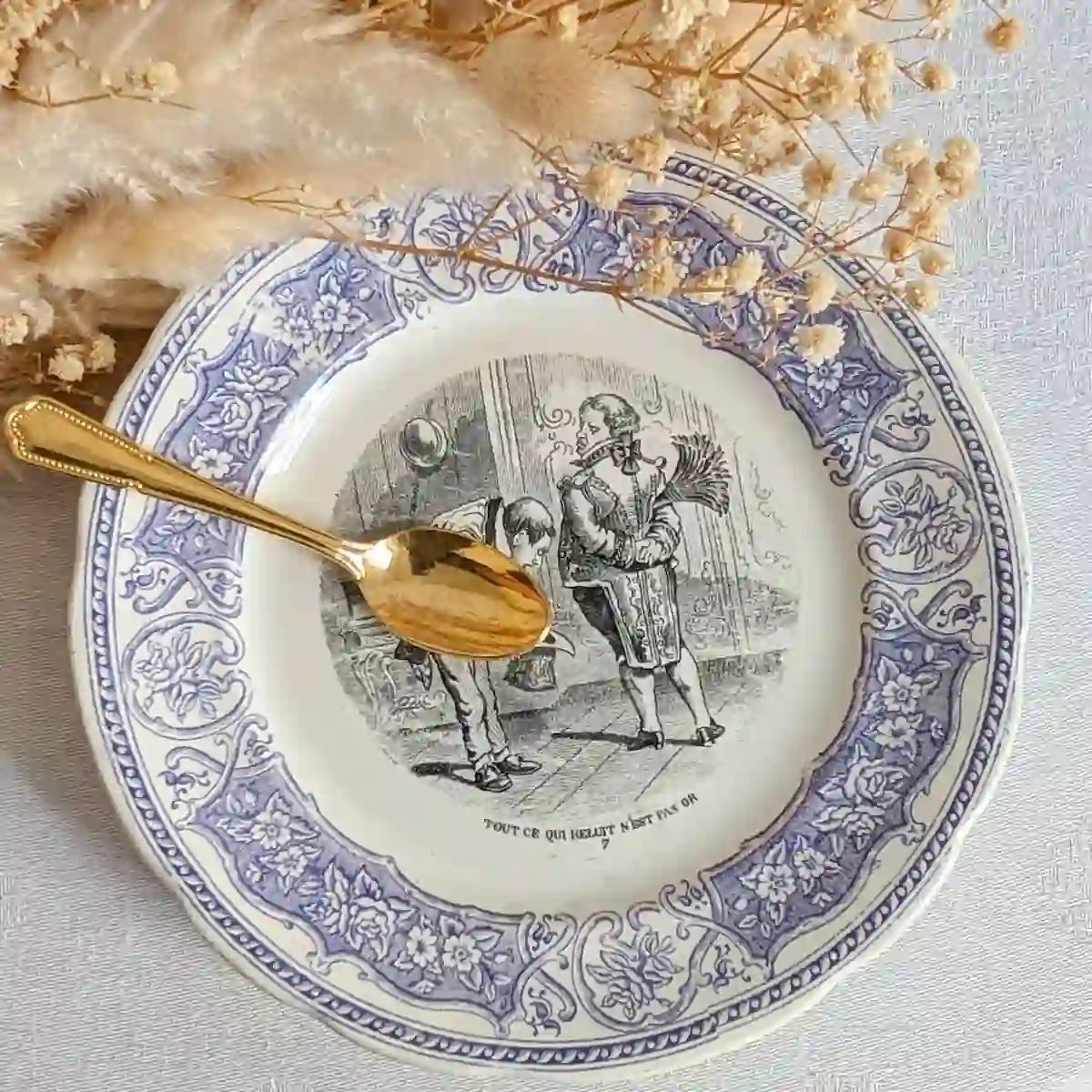 Assiette parlante Tout ce qui reluit n'est pas or, présentée avec une cuillère dorée et des fleurs séchées.