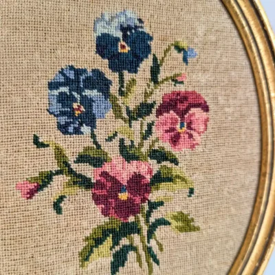 Détail des pensées bleues et roses brodées sur canevas beige, bordure dorée à droite.