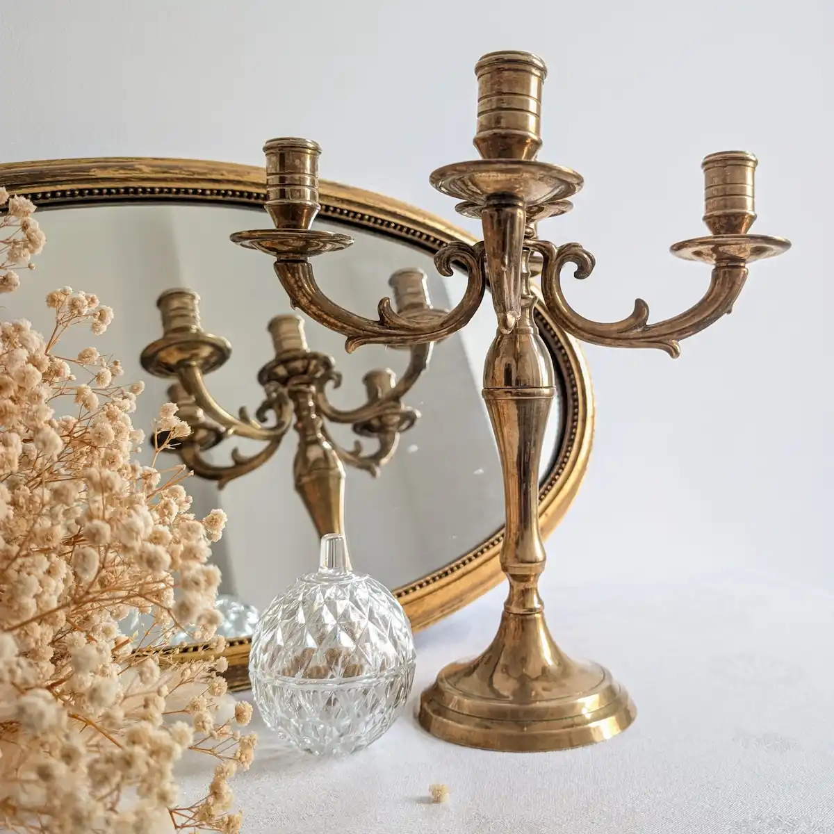 Chandelier en laiton devant un miroir ovale, à côté de fleurs séchées et d’un récipient en verre