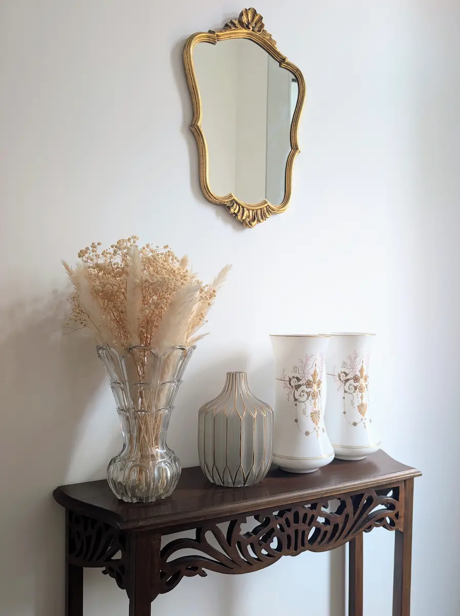 Miroir ancien à cadre doré au-dessus d'une console en bois décorée de vases vintage et de fleurs séchées