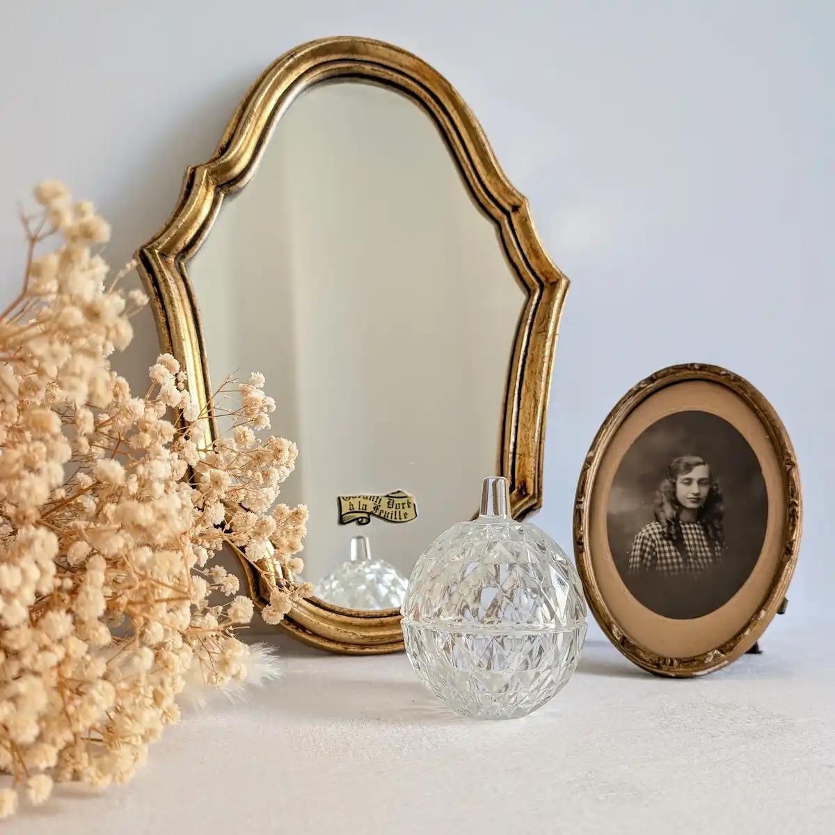 Miroir doré associé à des fleurs séchées, une boîte en verre et un portrait dans un cadre ovale.