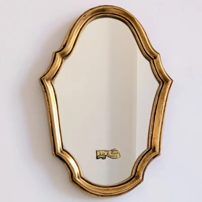 Petit miroir doré vu de face, avec un contour chantourné, une gorge sombre et une étiquette sur la glace.
