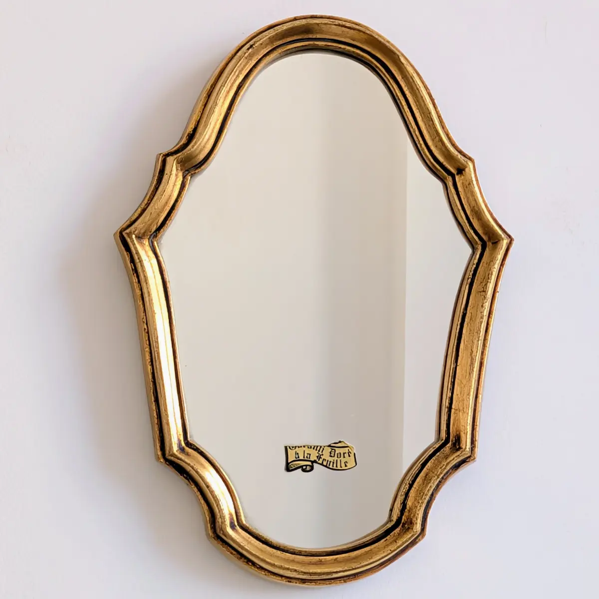 Petit miroir doré vu de face, avec un contour chantourné, une gorge sombre et une étiquette sur la glace.
