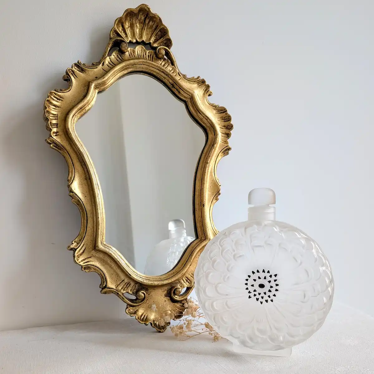 Petit miroir doré rocaille accroché au mur, avec un flacon décoratif blanc au premier plan.