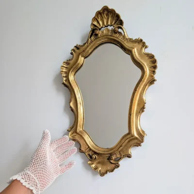 Petit miroir mural doré à coquilles présenté avec une main gantée pour montrer ses proportions.