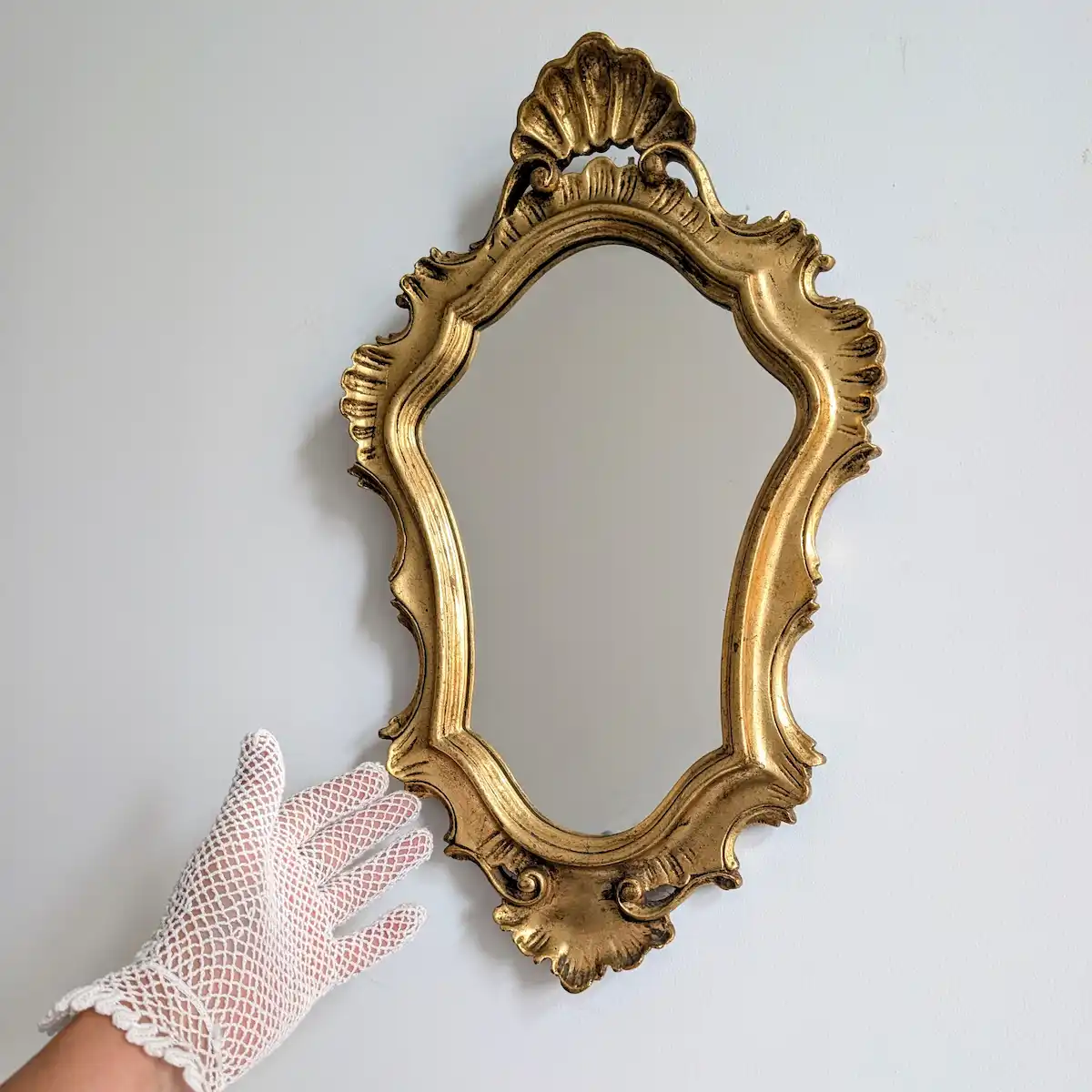 Petit miroir mural doré à coquilles présenté avec une main gantée pour montrer ses proportions.
