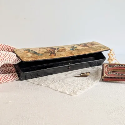 Plumier au couvercle entrouvert, montrant l’intérieur noir compartimenté et le petit fermoir en façade.