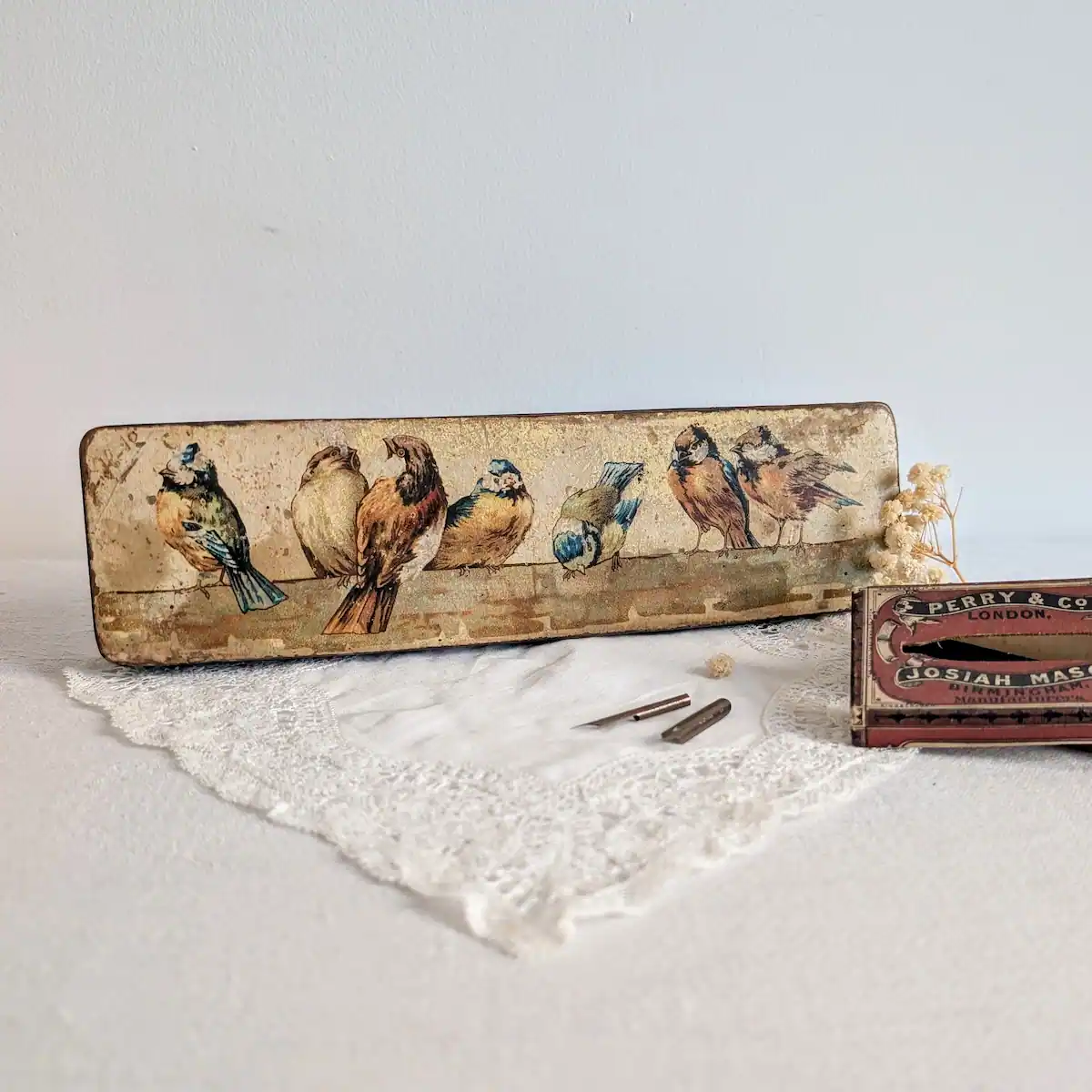Plumier à décor d’oiseaux présenté sur de la dentelle, entouré de plumes et d’une boîte d’accessoires.