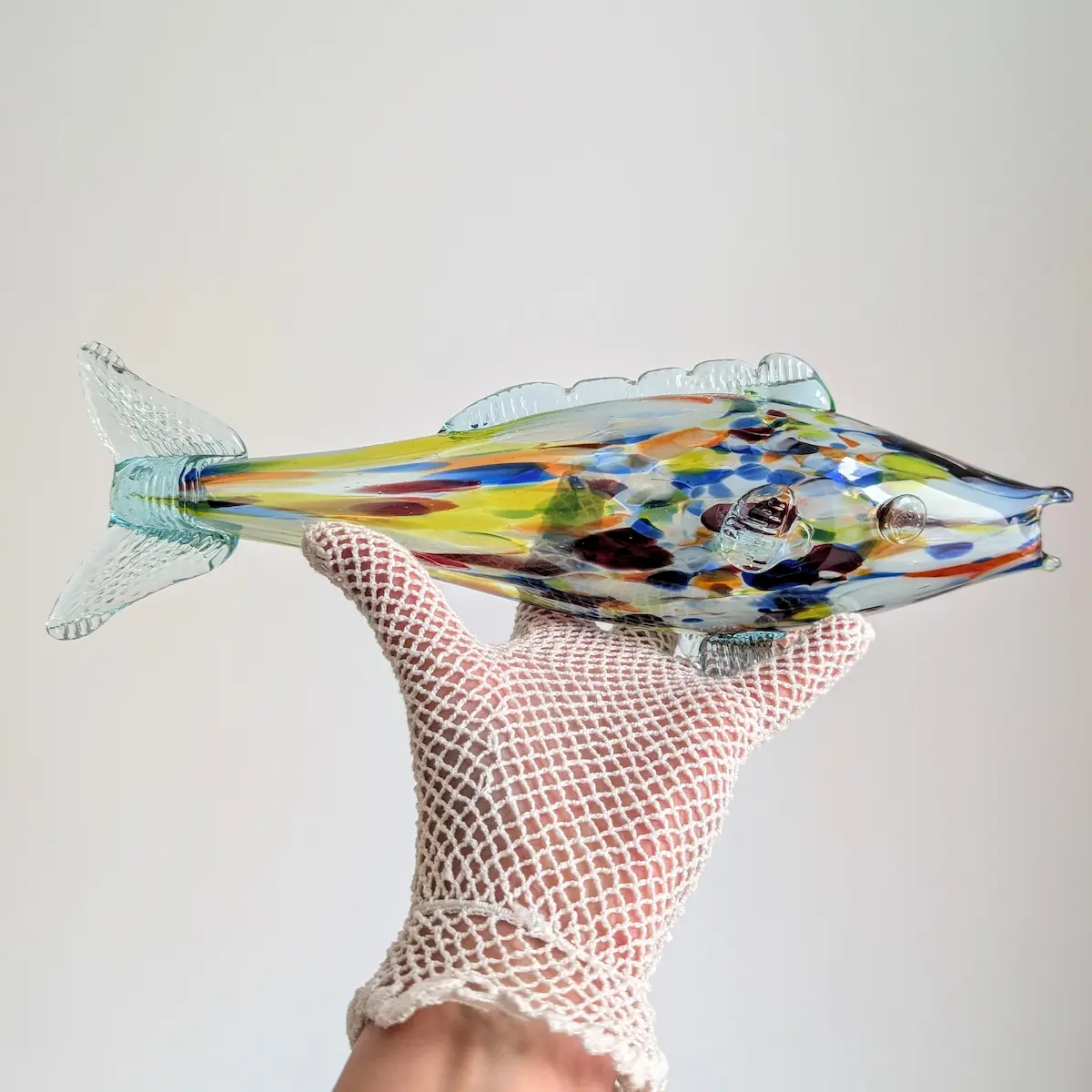 Poisson décoratif en verre multicolore présenté dans une main gantée pour apprécier ses proportions.