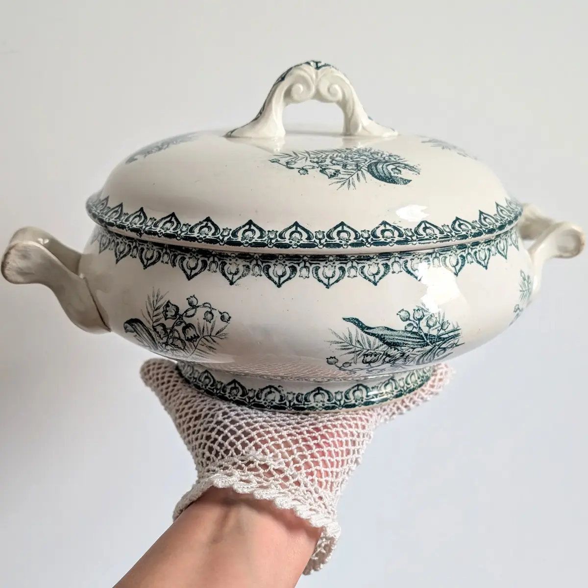 Soupière ancienne tenue sur une main gantée montrant ses deux anses, son piédouche et son décor vert.