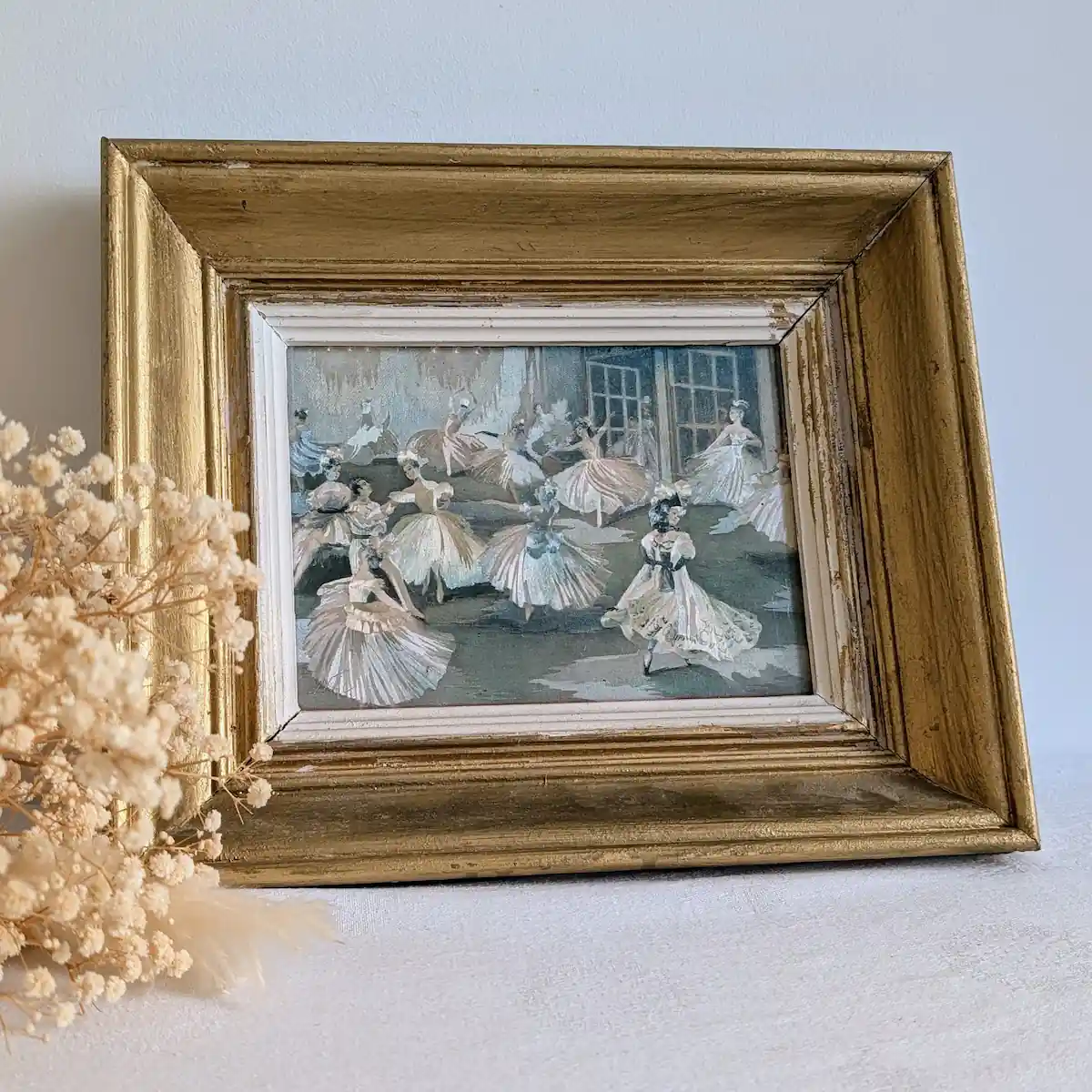 Tableau de ballet vintage encadré en bois doré, posé contre un mur clair à côté de fleurs séchées.